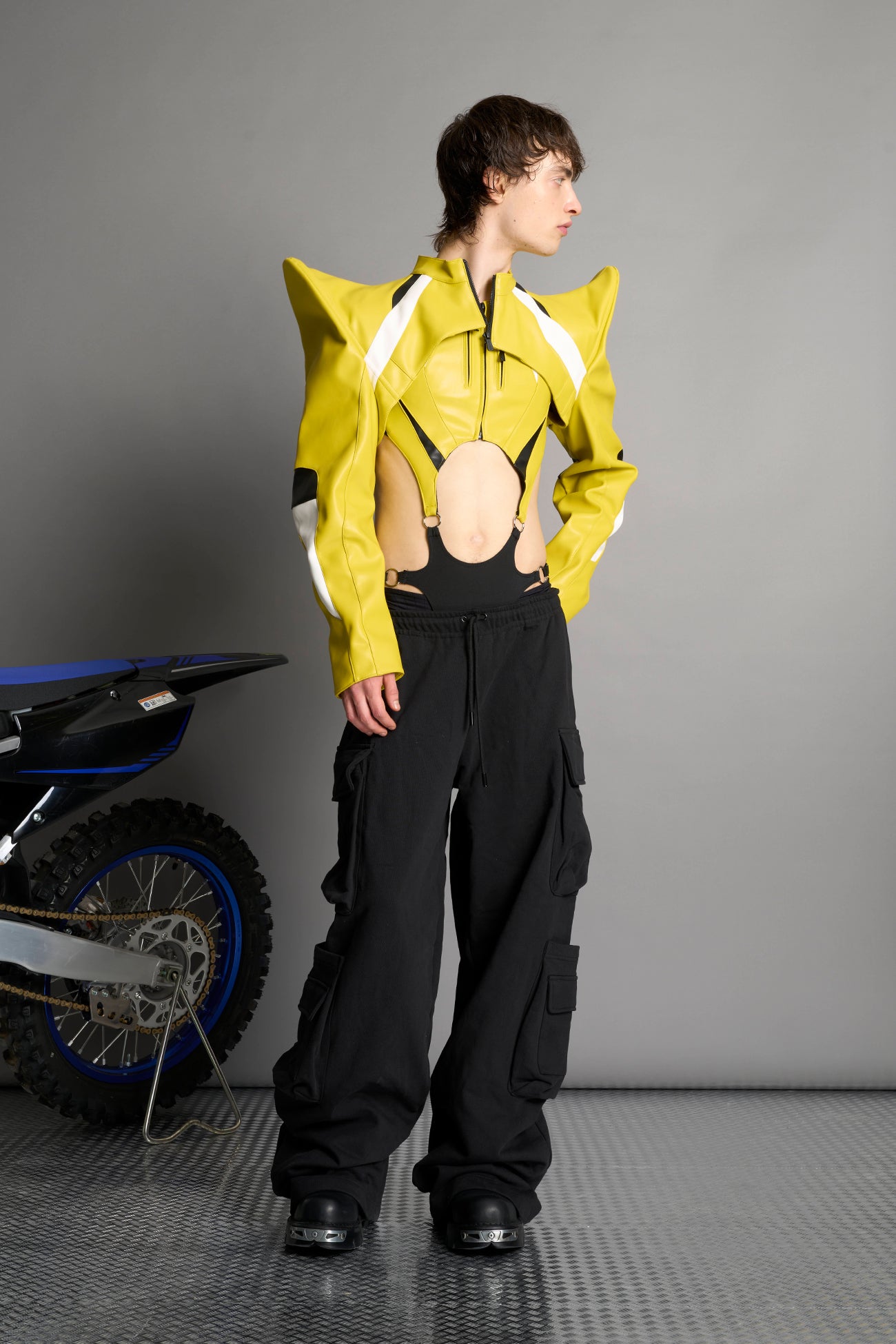 Model wears the yellow Namilia Spike Moto Bolero Jacket with wire shoulder pads, cropped hemline, branded front zipper, motocross-style lines, and black rubber logo tag on the back.