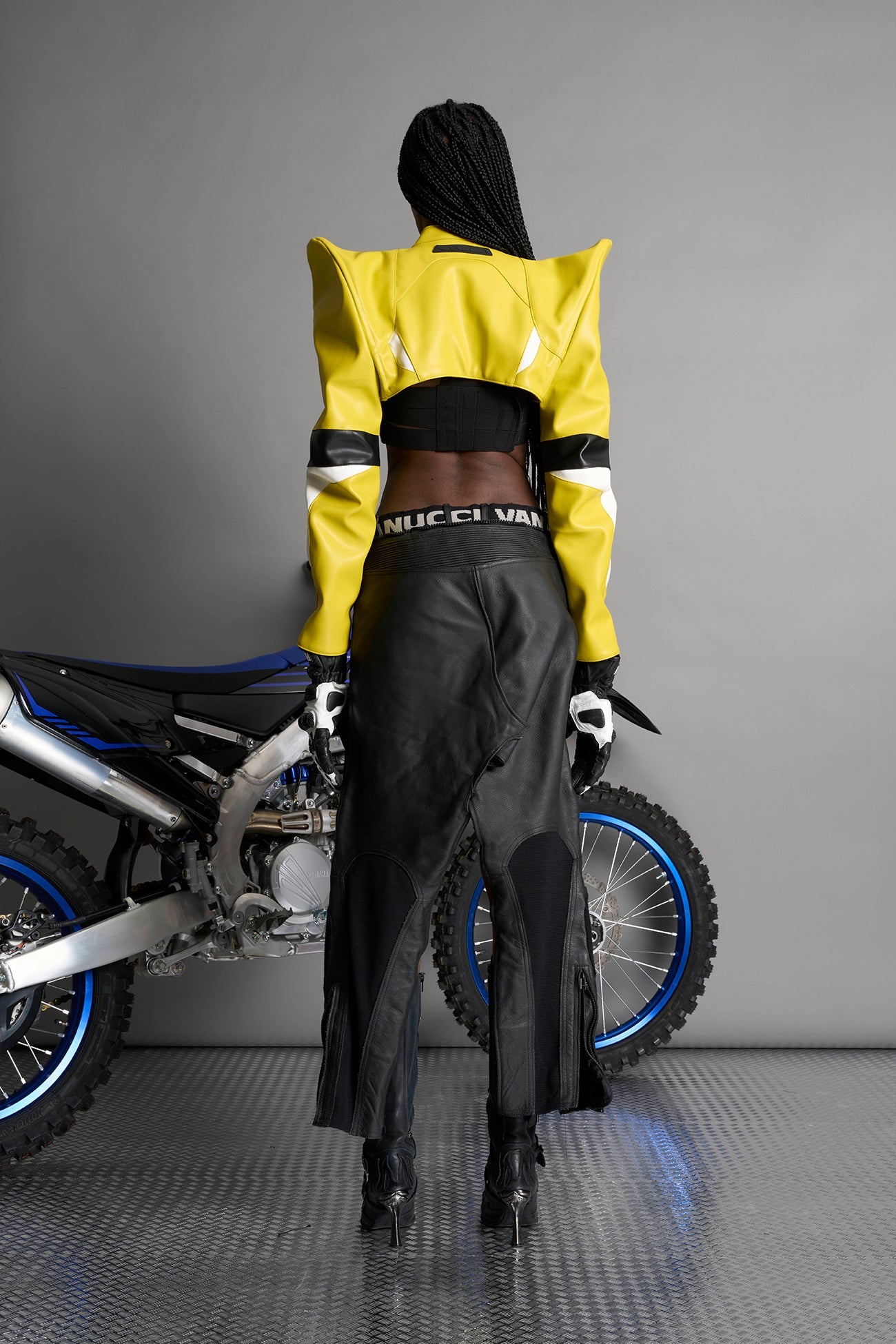 Model wears the yellow Namilia Spike Moto Bolero Jacket with wire shoulder pads, cropped hemline, branded front zipper, motocross-style lines, and black rubber logo tag on the back.