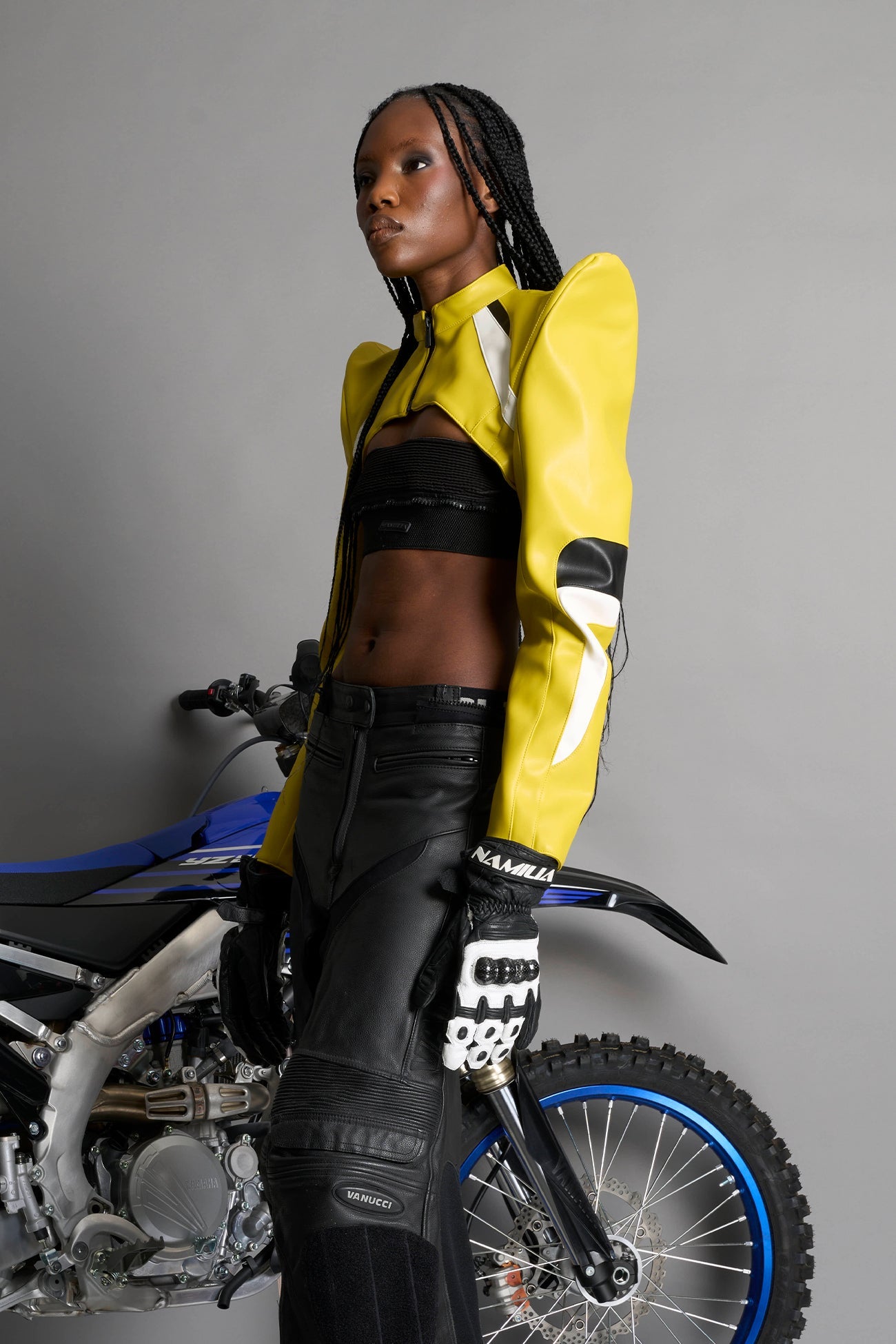 Model wears the yellow Namilia Spike Moto Bolero Jacket with wire shoulder pads, cropped hemline, branded front zipper, motocross-style lines, and black rubber logo tag on the back.