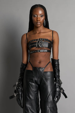 Black vegan leather belt top with three adjustable grommet straps, oversized metal buckles, and circular hardware – edgy bondage-inspired crop top for festival or clubwear.

