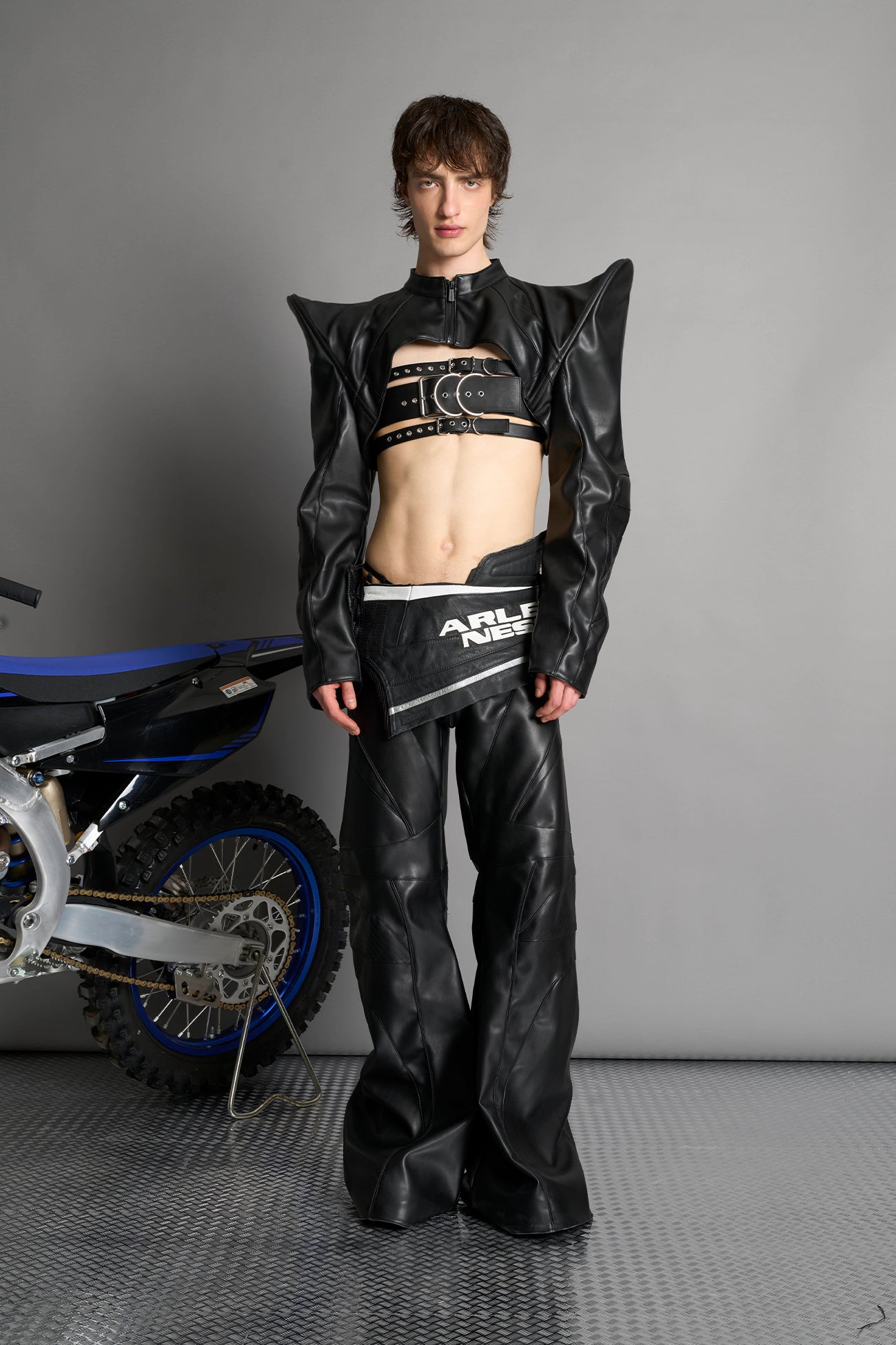 Model wears the black Namilia Spike Moto Bolero Jacket with wire shoulder pads, cropped hemline, branded front zipper, motocross-style lines, and black rubber logo tag on the back.