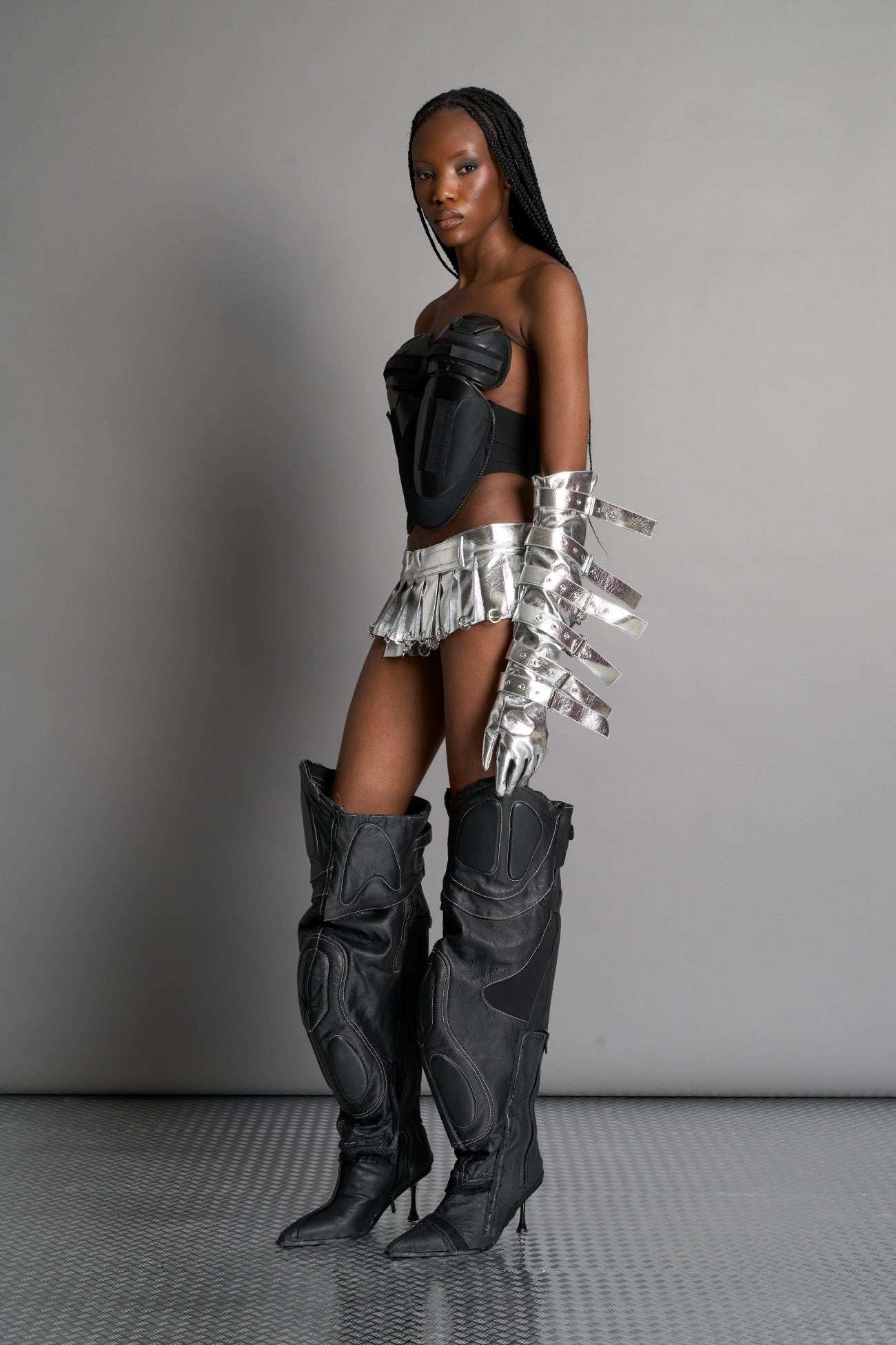 model wears the silver vegan PU leather Namilia Pleated Mini Skirt with ring piercing details on the hemline and velcro fold-over waistband closure.