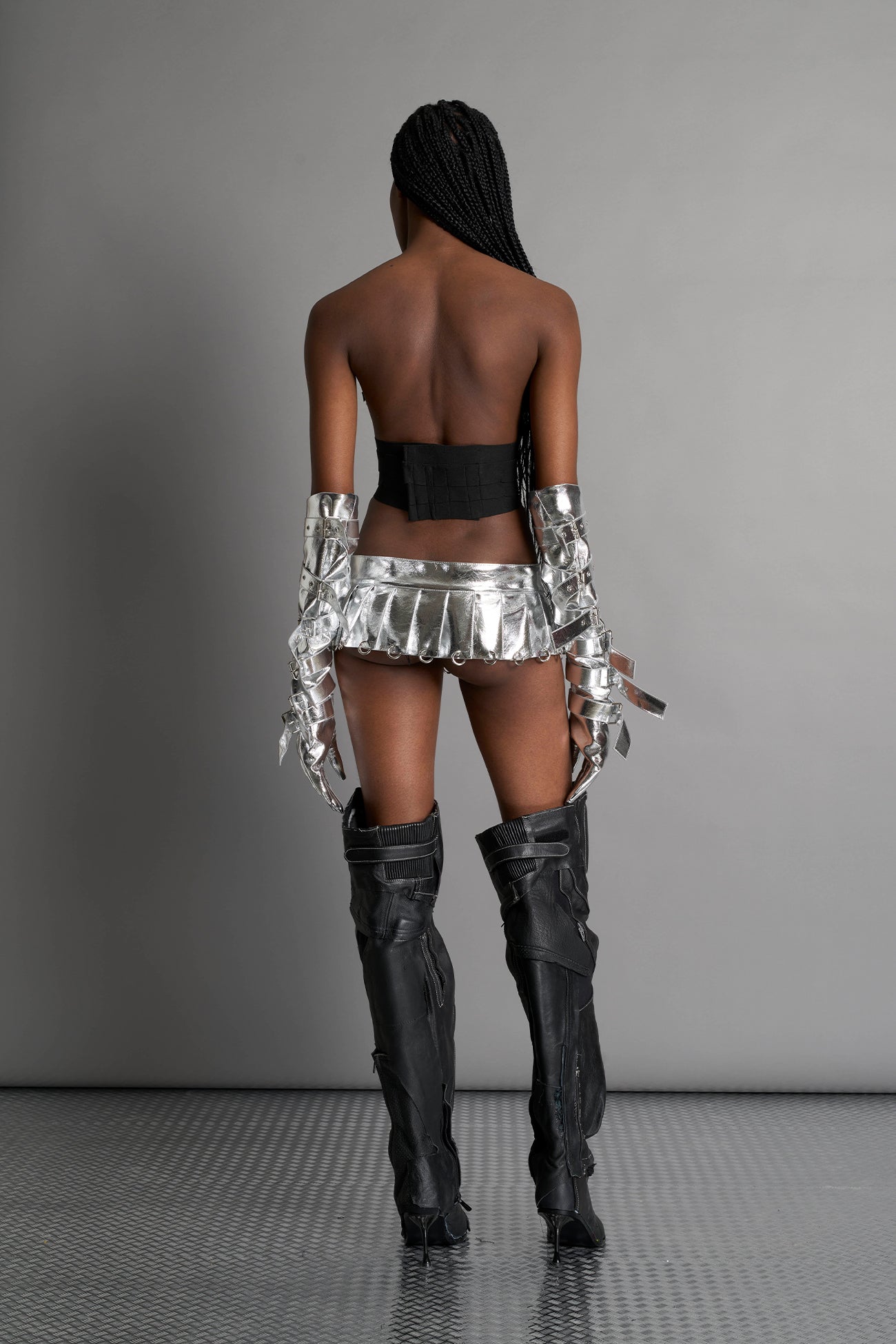 model wears the silver vegan PU leather Namilia Pleated Mini Skirt with ring piercing details on the hemline and velcro fold-over waistband closure.