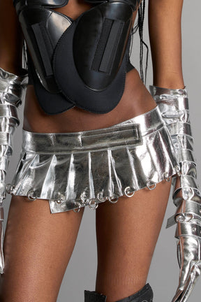 model wears the silver vegan PU leather Namilia Pleated Mini Skirt with ring piercing details on the hemline and velcro fold-over waistband closure.