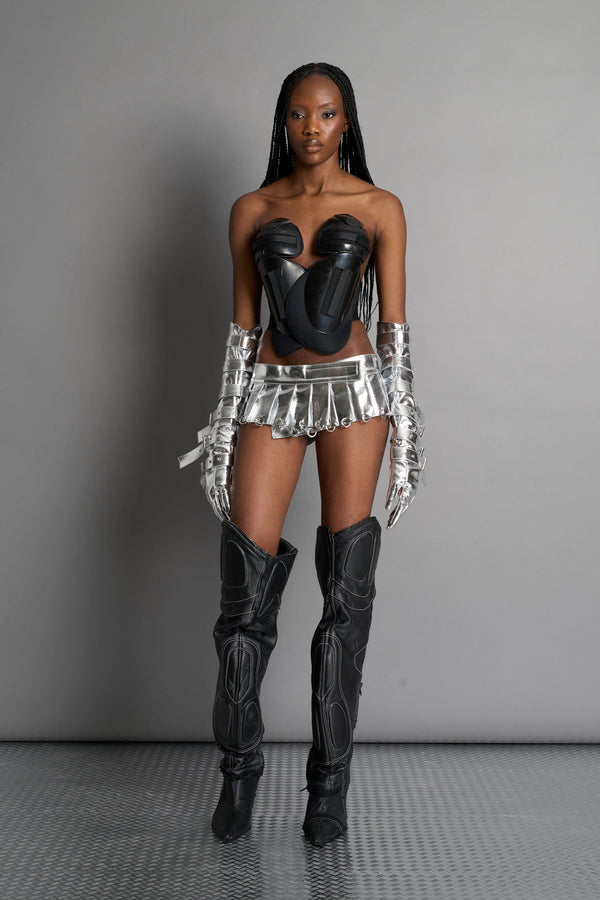 model wears the silver vegan PU leather Namilia Pleated Mini Skirt with ring piercing details on the hemline and velcro fold-over waistband closure. - SILVER