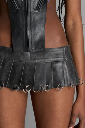 model wears the washed black vegan PU leather Namilia Pleated Mini Skirt with ring piercing details on the hemline and velcro fold-over waistband closure.