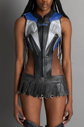 model wears the washed black vegan PU leather Namilia Pleated Mini Skirt with ring piercing details on the hemline and velcro fold-over waistband closure.