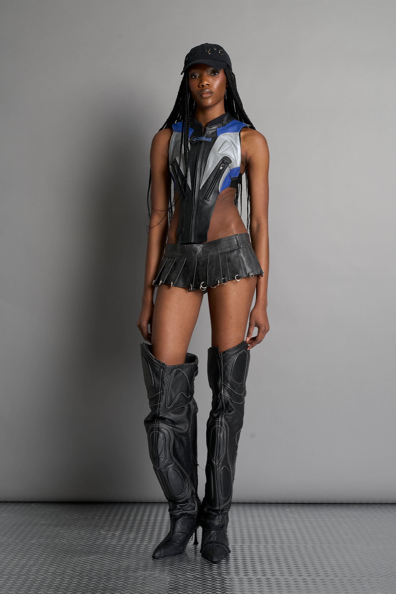 model wears the washed black vegan PU leather Namilia Pleated Mini Skirt with ring piercing details on the hemline and velcro fold-over waistband closure.