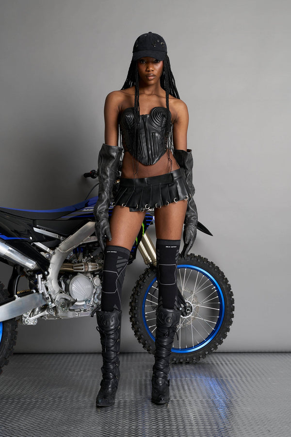 model wears the black Namilia Pleated Mini Skirt with ring piercing details on the hemline and velcro fold-over waistband closure. - BLACK