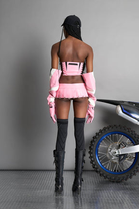 model wears the pink vegan PU leather Namilia Pleated Mini Skirt with ring piercing details on the hemline and velcro fold-over waistband closure.