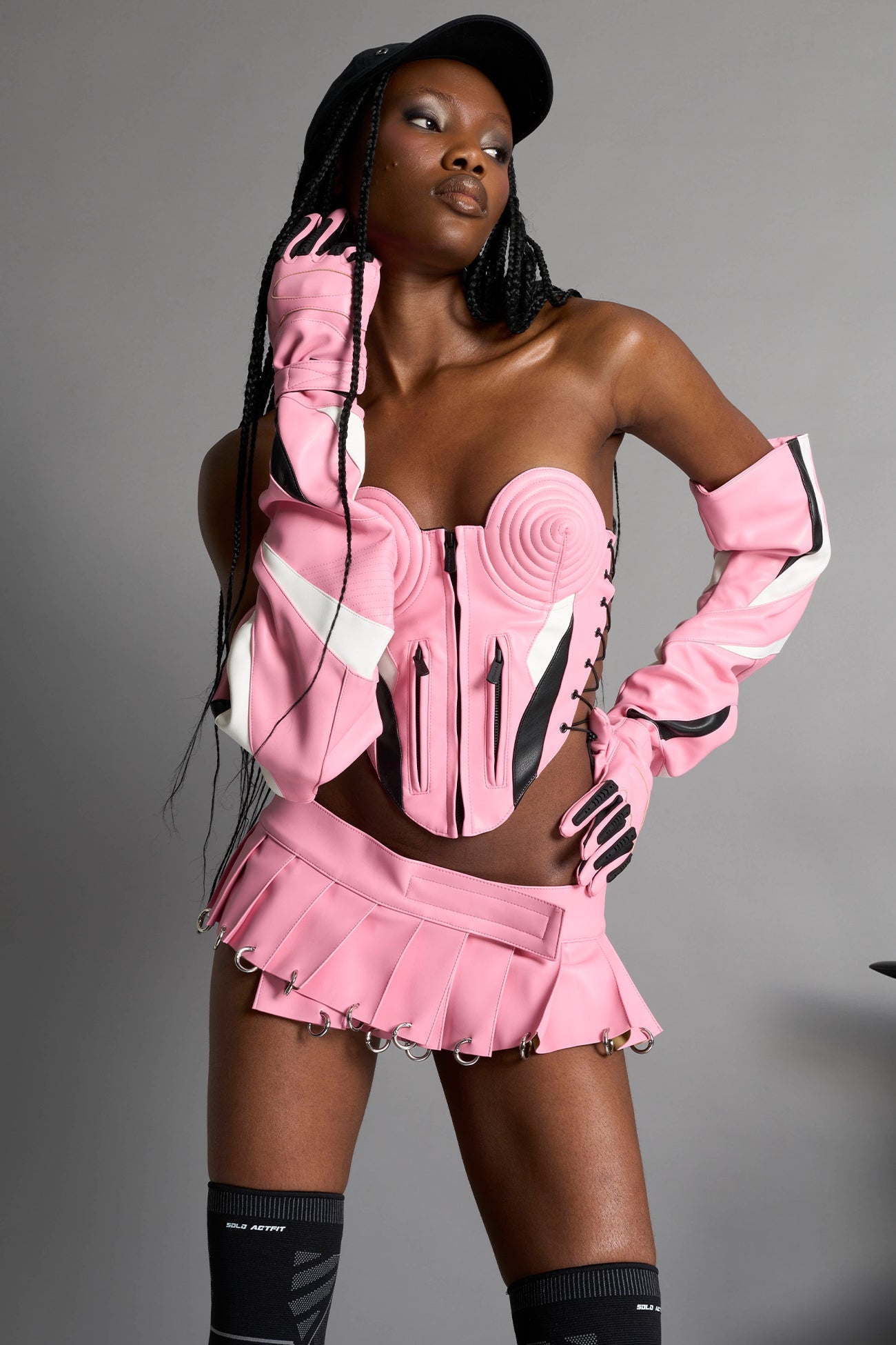model wears the pink vegan PU leather Namilia Pleated Mini Skirt with ring piercing details on the hemline and velcro fold-over waistband closure.