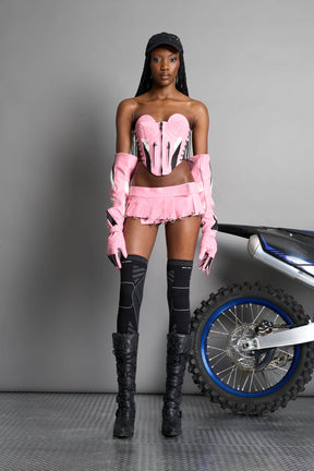 model wears the pink vegan PU leather Namilia Pleated Mini Skirt with ring piercing details on the hemline and velcro fold-over waistband closure.