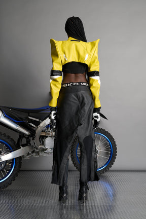 Model wears the yellow Namilia Spike Moto Bolero Jacket with wire shoulder pads, cropped hemline, branded front zipper, motocross-style lines, and black rubber logo tag on the back.