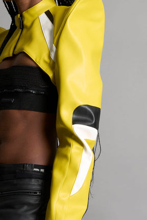 Model wears the yellow Namilia Spike Moto Bolero Jacket with wire shoulder pads, cropped hemline, branded front zipper, motocross-style lines, and black rubber logo tag on the back.