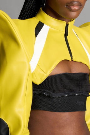 Model wears the yellow Namilia Spike Moto Bolero Jacket with wire shoulder pads, cropped hemline, branded front zipper, motocross-style lines, and black rubber logo tag on the back.