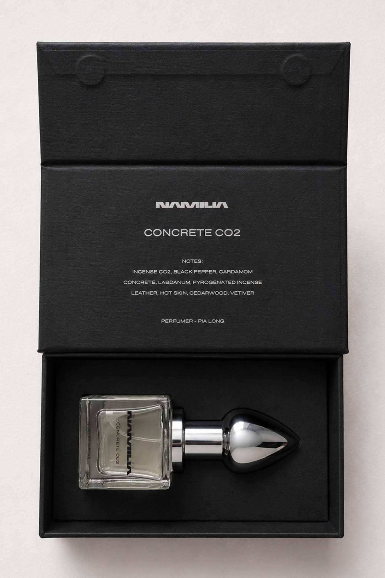 A perfume bottle with a clear rectangular glass body and a metallic silver dropper cap, presented inside an open black box. The inside lid shows the brand name 'NAMILIA' and the scent name 'CONCRETE CO2'.