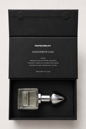 A perfume bottle with a clear rectangular glass body and a metallic silver dropper cap, presented inside an open black box. The inside lid shows the brand name 'NAMILIA' and the scent name 'CONCRETE CO2'.