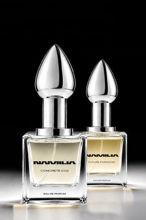 Two Namila perfume bottles with metallic silver teardrop-shaped caps and clear glass bases, filled with pale yellow liquid, set against a solid black background.